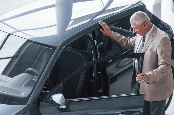 A imagem mostra um homem mais velho, de cabelos grisalhos, vestindo um terno bege e gravata, interagindo com um carro estacionado em um ambiente fechado. Ele está com uma das mãos apoiada na porta aberta do carro, olhando para o interior do veículo. O carro parece ser moderno e de cor escura.