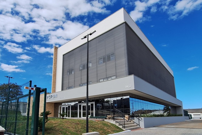 A imagem mostra um edifício moderno, identificado como um fórum. A construção tem linhas retas e fachadas envidraçadas, com tons de branco e cinza. Há uma entrada com escadas e um caminho pavimentado à frente, além de um gramado bem cuidado. O céu está azul com algumas nuvens, proporcionando um cenário claro e arejado.
