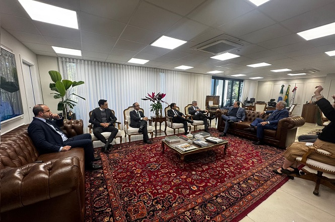 A imagem mostra uma sala de reunião com várias pessoas sentadas em sofás e cadeiras dispostas em círculos. A sala possui um tapete vermelho decorativo no chão, mesas de centro com revistas e livros, e algumas plantas decorativas. Há cortinas nas janelas e as paredes têm quadros pendurados. O ambiente parece ser formal, possivelmente um escritório ou uma área de reuniões. As pessoas estão vestidas de maneira formal e conversam entre si. É um espaço bem iluminado com luzes no teto. Há também bandeiras no canto da sala.