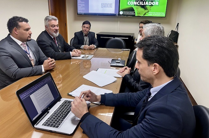 A imagem mostra um grupo de seis homens sentados ao redor de uma mesa de reunião. Eles estão em uma sala de conferências, onde há duas telas ao fundo com informações exibidas. Na mesa, há um laptop aberto, alguns papéis e documentos. Todos estão vestidos formalmente, usando ternos e gravatas, sugerindo um encontro de negócios ou discussão profissional. Eles estão engajados em uma conversa, indicando que uma reunião ou discussão está ocorrendo.