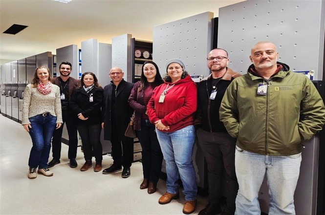 A imagem mostra um grupo de nove pessoas posando para uma foto em um ambiente interno. Elas estão alinhadas em frente a um conjunto de armários ou estantes grandes, possivelmente um ambiente de trabalho ou arquivo. As pessoas estão vestindo roupas de inverno, como jaquetas e casacos, e algumas delas estão sorrindo para a câmera. A iluminação é clara e o chão parece ser de material liso.