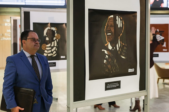 Na imagem, há um homem de terno azul olhando para uma exposição de arte. Ele está segurando um objeto que parece uma pasta ou portfólio. Em frente a ele, há um quadro em exibição com uma imagem artística de uma pessoa sorrindo. O fundo da imagem mostra outras obras de arte e pessoas na galeria. A cena transmite um ambiente de evento ou exibição cultural.