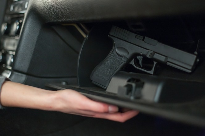 A imagem mostra uma mão abrindo um compartimento no interior de um carro, revelando uma pistola guardada dentro. O foco está no armamento, e o fundo está levemente desfocado.
