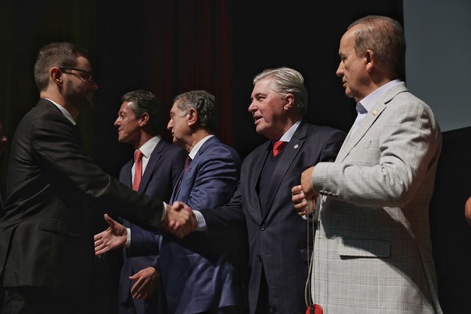 A imagem mostra um grupo de cinco homens, todos vestindo ternos. Eles estão em pé, aparentemente em um evento formal. Um dos homens está apertando a mão de outro. A cena parece ser uma cerimônia ou um encontro oficial. O fundo é escuro, e a iluminação está focada nos homens.