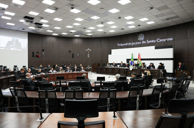 A imagem mostra uma sala de tribunal no Tribunal de Justiça de Santa Catarina. Há várias pessoas sentadas ao redor de uma grande mesa em forma de "U", participando de uma sessão. Na parede ao fundo, estão as palavras "Tribunal de Justiça de Santa Catarina", além de um brasão. Há bandeiras colocadas em destaque, incluindo a bandeira do Brasil e de Santa Catarina. O ambiente é iluminado com um teto repleto de luzes e há cadeiras adicionais e equipamentos audiovisuais ao redor.