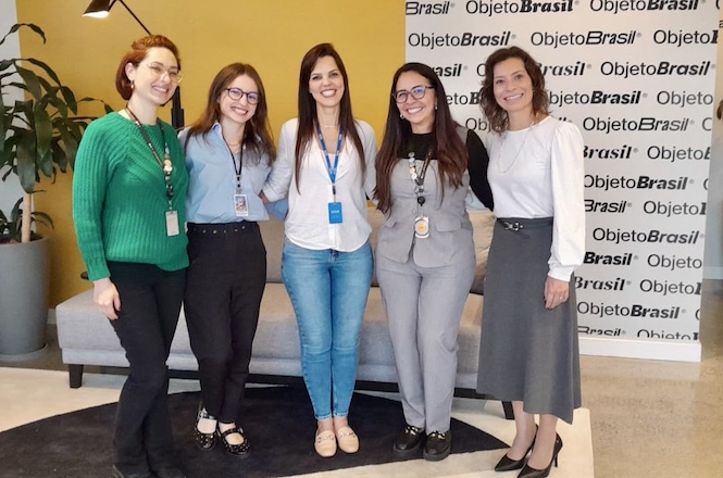 A imagem mostra um grupo de pessoas em uma sala que parece ser uma biblioteca, com estantes cheias de livros ao fundo. No centro, algumas pessoas estão em pé, enquanto outras, vestidas de azul, estão sentadas em bancos, provavelmente participando de uma reunião ou apresentação. A sala tem janelas altas, permitindo a entrada de luz natural.