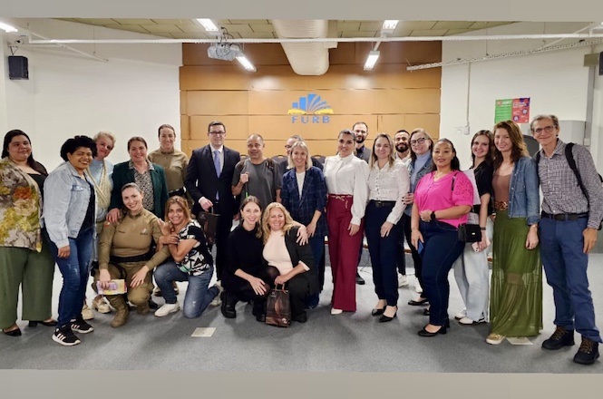 A imagem mostra um grupo de pessoas posando para uma foto em um ambiente interno. Algumas pessoas estão vestindo roupas formais, enquanto outras estão vestidas de forma mais casual. Ao fundo, há uma parede com o logo da "FURB". O chão é coberto por carpete cinza e no teto há iluminação branca. As pessoas parecem estar em um tipo de evento ou reunião institucional.