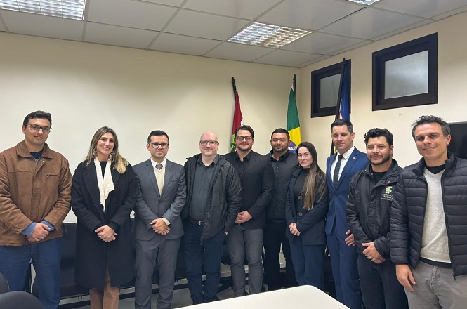A imagem mostra um grupo de onze pessoas posando para uma foto em um ambiente interno. Ao fundo, há três bandeiras em mastros. As pessoas estão vestidas formalmente, algumas com ternos e outras com roupas de frio. Elas estão alinhadas lado a lado, sorrindo para a câmera. Parece ser um evento oficial ou reunião, dada a presença das bandeiras.