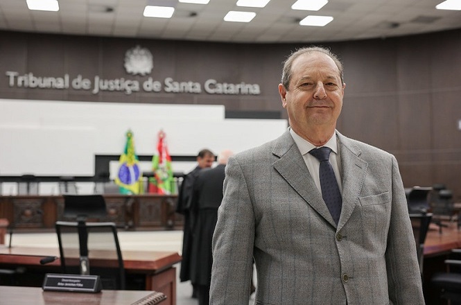 A imagem mostra um ambiente formal de tribunal, especificamente o Tribunal de Justiça de Santa Catarina. Em primeiro plano, há um homem vestido com terno e gravata, aparentando estar em destaque. Ao fundo, veem-se bandeiras e outras pessoas também em trajes formais, possivelmente em atividade judicial ou solenidade. O local tem um aspecto organizado e oficial, com móveis e decoração adequados a um tribunal.