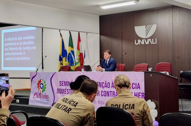 A imagem mostra um evento em uma sala de conferências com várias pessoas assistindo. Há uma mesa com um palestrante sentado, e atrás dele estão bandeiras e um logotipo da UNIVALI na parede. Um telão exibe uma apresentação cujo tema está relacionado à eficácia de medidas protetivas e desafios do sistema de justiça na luta contra a violência doméstica. Na frente, um banner roxo menciona um seminário sobre prevenção à violência doméstica e contra a mulher em Itajaí. Participantes da Polícia Militar estão na plateia.