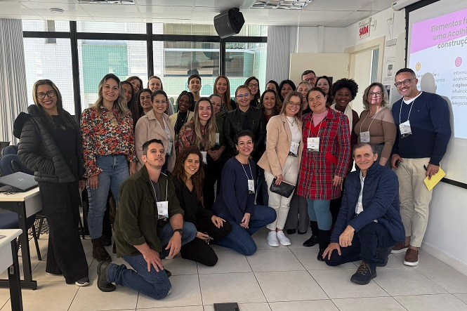 A imagem mostra um grupo de pessoas posando para uma foto em uma sala de aula ou conferência. Todos estão sorrindo e muitos vestem crachás. Ao fundo, há uma tela de projeção com um slide visível. A atmosfera parece descontraída e profissional.