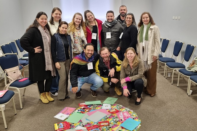 A imagem mostra um grupo de treze pessoas posando para a foto em uma sala. Elas estão organizadas em duas filas, sendo que algumas estão agachadas na frente. O ambiente parece ser uma sala de reuniões ou um espaço para workshops, com cadeiras azuis organizadas contra as paredes. No centro do grupo, no chão, há vários papéis coloridos dispostos em círculo. As pessoas na imagem estão sorrindo e parecem felizes.