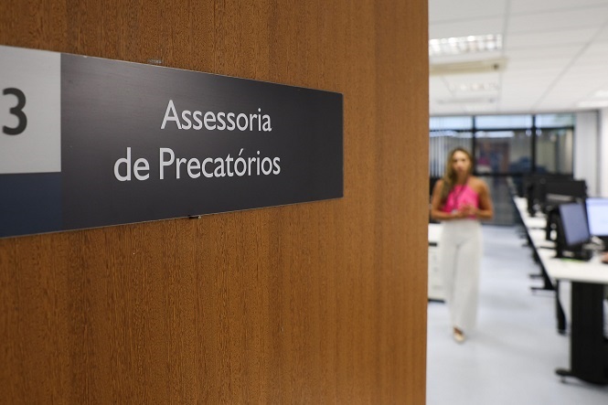 A imagem mostra a entrada de uma sala com uma placa na porta indicando "Assessoria de Precatórios". A porta está aberta, revelando parte de um escritório ao fundo, onde várias mesas e computadores estão dispostos. Há uma pessoa em pé, usando roupas formais, com um vestido rosa e calças brancas, dentro do escritório. A decoração do ambiente é moderna, com iluminação de teto em um ambiente bem iluminado.