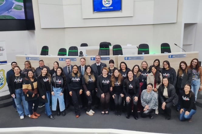 A imagem mostra um grupo de pessoas posando para uma foto em um auditório. Ao fundo, há várias cadeiras e mesas, provavelmente para debates ou apresentações. As pessoas na frente estão sorrindo e muitas delas vestem camisetas pretas com uma inscrição. O ambiente parece ser formal, possivelmente um local governamental ou instituição pública, devido ao painel e emblema visíveis nas paredes.