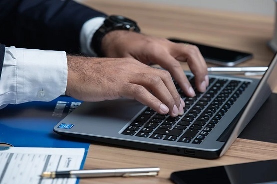 A imagem mostra um homem usando um laptop sobre uma mesa. Suas mãos estão digitando no teclado. Ao lado do laptop, há um smartphone, um caderno ou documento com um clipe, e uma caneta sobre a mesa. A pessoa parece estar vestindo um terno escuro. O ambiente parece ser um espaço de escritório.