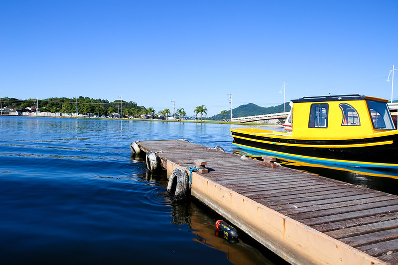 A imagem mostra um píer sobre uma água calma, com um barco amarelo e preto próximo ao canto direito. O céu é azul e claro. Ao fundo, vê-se uma linha costeira com algumas palmeiras, outros elementos de vegetação e algumas construções. Existem também algumas montanhas ao longe. A cena parece ser um dia ensolarado e tranquilo à beira-mar ou à beira de um lago.
