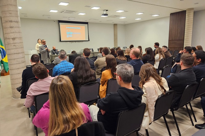 A imagem mostra uma sala de conferências com várias pessoas sentadas, assistindo a uma apresentação. Na frente da sala, duas pessoas estão em pé falando para o público, enquanto uma tela de projeção exibe um slide. Algumas bandeiras estão posicionadas à esquerda, e o ambiente é bem iluminado, com teto rebaixado e paredes claras.