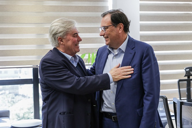 A imagem mostra dois homens vestindo ternos, sorrindo e cumprimentando-se de forma amigável em um ambiente interno. Eles estão frente a frente, com um dos homens colocando a mão no peito do outro. Ao fundo, é possível ver uma janela com persianas e algumas cadeiras. A atmosfera parece ser de camaradagem ou encontro profissional