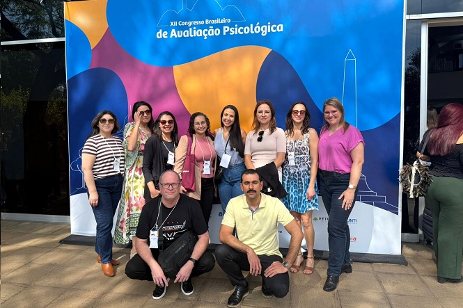 A imagem mostra um grupo de pessoas posando para uma foto em frente a um painel colorido. O painel é do "XII Congresso Brasileiro de Avaliação Psicológica". As pessoas parecem estar em clima de confraternização, algumas delas usando crachás. O fundo do painel exibe cores vibrantes, como azul, laranja, rosa e roxo, e na parte superior do painel há o logotipo do evento. A atmosfera parece ser de um evento profissional ou acadêmico.