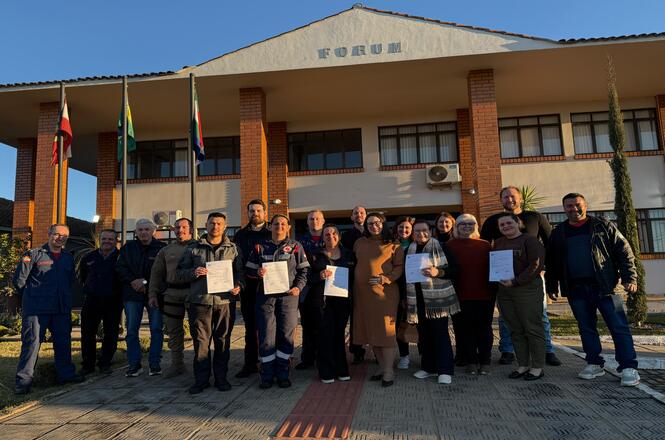 A imagem mostra um grupo de pessoas posando em frente a um edifício identificado como "FÓRUM". O grupo está em uma área externa, próximo a algumas bandeiras. Algumas pessoas seguram certificados ou documentos. O ambiente parece ser agradável, com o sol iluminando a cena.