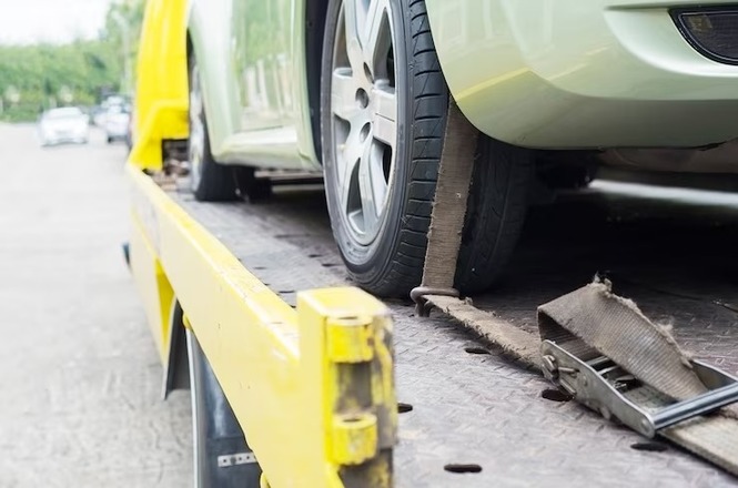 A imagem mostra um carro verde sobre um caminhão guincho amarelo. A roda dianteira do carro está fixada com uma correia de segurança, e o caminhão guincho está em uma estrada ou estacionamento, com algumas árvores visíveis ao fundo.