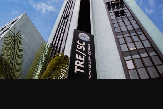A imagem mostra um edifício alto com janelas de vidro espelhado, visto de baixo para cima. Na frente do prédio, há uma placa com o logo e a sigla "TRE/SC", que se refere ao Tribunal Regional Eleitoral de Santa Catarina. Ao lado da placa, parte de uma palmeira é visível. O céu está azul ao fundo.