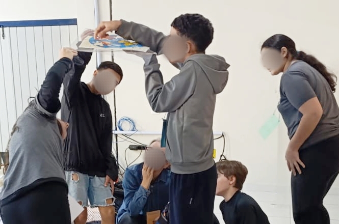Na imagem, há um grupo de pessoas interagindo. Uma pessoa está segurando um cartaz colorido, enquanto outras estão em pé ou agachadas ao redor, parecendo concentradas na atividade. O ambiente tem paredes claras e alguns fios visíveis no fundo. O grupo parece estar envolvido em uma atividade colaborativa ou educacional.