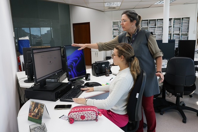 A imagem mostra duas mulheres em um ambiente de escritório. A mulher sentada está usando um computador enquanto a mulher em pé está ao seu lado, apontando para a tela. A mesa está coberta por itens como um estojo, um celular e um calendário. Ao fundo, há prateleiras cheias de documentos e mais equipamentos de escritório. A cena retrata um ambiente de trabalho colaborativo.
