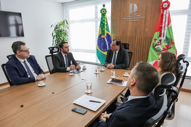 Foto horizontal: Francisco Oliveira Neto entre as bandeiras do Brasil e de SC participa de reunião com autoridades, tons claros.