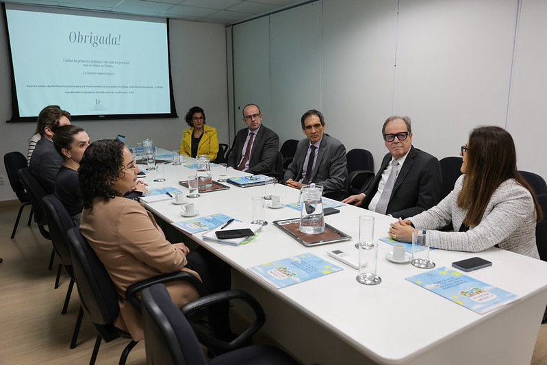 A imagem mostra um grupo de pessoas sentadas ao redor de uma mesa de reunião em uma sala de conferências. Há oito pessoas, sendo cinco de um lado e três do outro, todas vestidas formalmente. Na mesa, há materiais de papelaria, copos, garrafas e xícaras de café. Ao fundo, há uma tela de apresentação exibindo a palavra "Obrigada!", que indica um provável contexto profissional ou empresarial. As pessoas parecem estar engajadas em uma discussão ou apresentação.