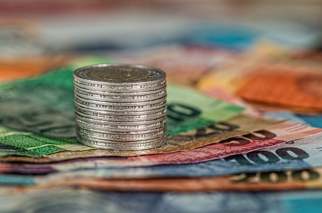 A imagem mostra uma pilha de moedas de metal prateado em primeiro plano, com várias notas de dinheiro coloridas espalhadas ao redor. A composição sugere um tema relacionado a finanças ou economia, destacando o contraste entre moedas e cédulas. As cores das notas variam, incluindo verde, azul e laranja.