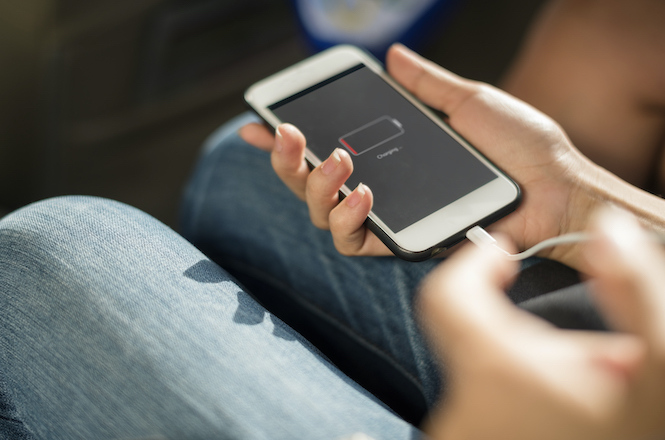A imagem mostra uma pessoa segurando um smartphone nas mãos. A tela do aparelho exibe um ícone de bateria descarregada, indicando que o telefone precisa ser carregado. O cabo de carregamento está conectado ao dispositivo. A pessoa está vestindo jeans, e o ambiente parece ser casual e descontraído.