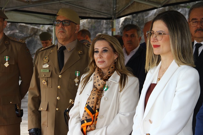 A imagem mostra um grupo de pessoas em um evento formal. À esquerda, há homens em uniformes militares com medalhas, e ao lado, estão duas mulheres. A mulher do meio veste um casaco claro e um lenço estampado, e a mulher à direita usa óculos e um blazer branco. O cenário parece ser ao ar livre e coberto por uma tenda.