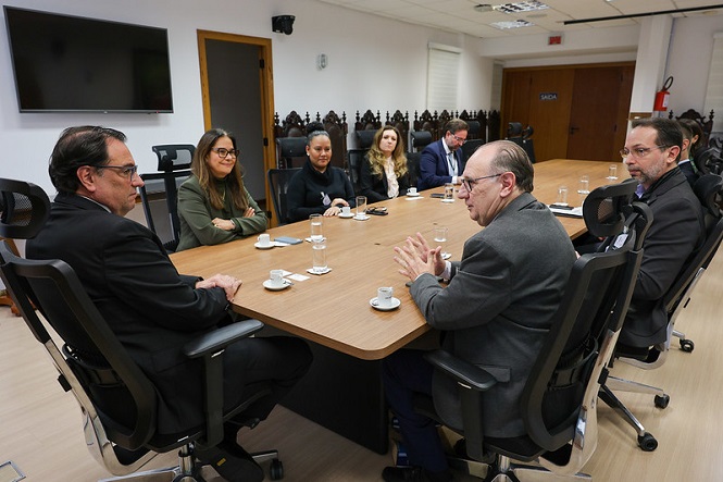A imagem mostra um grupo de pessoas sentadas ao redor de uma mesa de reunião. Há sete pessoas visíveis, algumas vestindo roupas formais. Na mesa, há copos de água e xícaras de café. A sala parece ser um espaço de escritório com cadeiras estofadas em volta da mesa, portas de madeira e uma tela na parede. A atmosfera sugere uma reunião ou discussão profissional.