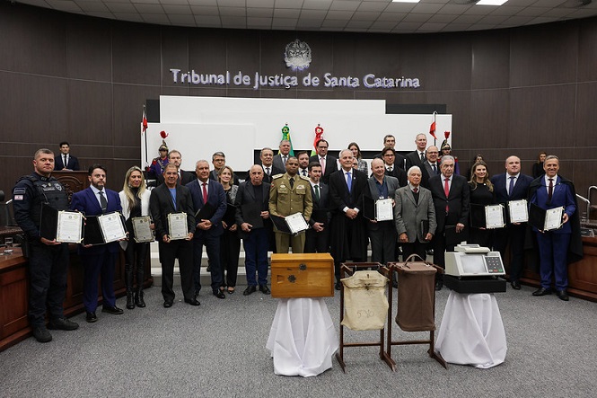 A imagem mostra um grupo de pessoas posando para uma foto em um ambiente formal. Elas estão dentro de uma sala do Tribunal de Justiça de Santa Catarina. Algumas pessoas seguram certificados ou prêmios. No centro, há objetos em exibição sobre mesas cobertas com toalhas brancas. O local tem um ambiente sério, com uma decoração formal, e é possível ver bandeiras ao fundo. O grupo é composto por homens e mulheres vestidos formalmente.