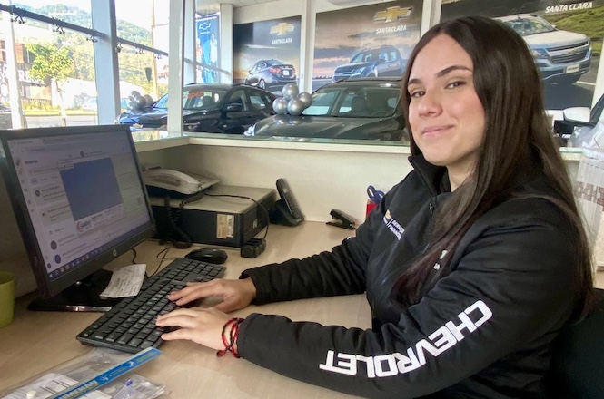 Na imagem, há uma mulher sentada em uma mesa de trabalho. Ela está usando um casaco preto com a palavra "CHEVROLET" no braço e está sorrindo. Em frente a ela, há um computador sobre a mesa. Ao fundo, vê-se o interior de uma concessionária de carros, com veículos e banners da Chevrolet. Através das janelas, algumas árvores e parte do exterior são visíveis. A cena sugere um ambiente de trabalho em uma concessionária de automóveis.