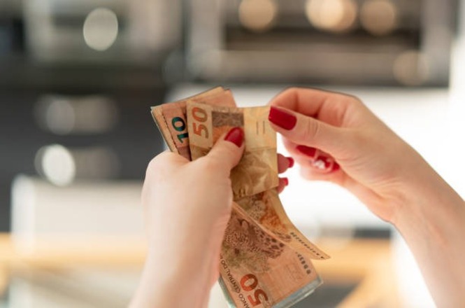 A imagem mostra as mãos de uma mulher contando notas de dinheiro. As notas são de reais, o que indica que é dinheiro brasileiro. A mulher usa esmalte vermelho nas unhas. O fundo da imagem está desfocado, destacando a ação de manusear o dinheiro.