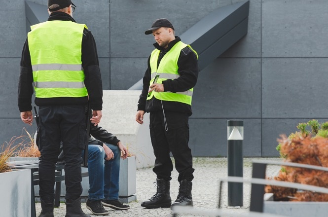 A imagem mostra dois homens de coletes refletivos amarelos, possivelmente seguranças, conversando com um homem sentado. Eles estão em uma área externa, próxima a um edifício cinza. Adicionalmente, há algumas plantas e luminárias na cena. Os seguranças estão vestidos com roupas escuras e um deles parece estar segurando um rádio comunicador.