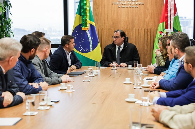 Foto horizontal: Francisco Oliveira Neto conduz reunião entre bandeiras do Brasil e SC em ambiente institucional.