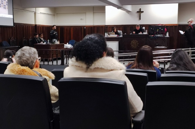 A imagem mostra uma sala de tribunal. Há várias pessoas sentadas em cadeiras, algumas usando casacos com gola de pelo. Na frente, há um juiz e outros funcionários em uma bancada elevada. Um crucifixo está pendurado na parede ao fundo. À esquerda, há um projetor de tela. O ambiente parece formal e sério, como é típico em um tribunal.