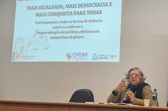 A imagem mostra uma mulher sentada atrás de uma mesa, falando em um microfone. Há uma apresentação projetada na parede atrás dela, com o título "MAIS IGUALDADE, MAIS DEMOCRACIA E MAIS CONQUISTA PARA TODAS". O texto menciona enfrentamento à violência contra as mulheres e a democratização das políticas públicas com perspectiva de gênero. Há logotipos de organizações e instituições na parte inferior da projeção. A mulher parece estar participando de um evento ou palestra relacionado ao tema.