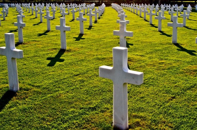 A imagem mostra um cemitério militar com várias cruzes brancas alinhadas em um gramado bem cuidado. As cruzes marcando os túmulos são dispostas em fileiras simétricas, criando um padrão visual impressionante. A luz do sol ilumina a cena, projetando sombras das cruzes sobre o gramado.