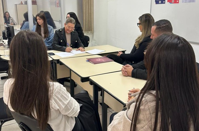 Na imagem, há um grupo de pessoas sentadas em torno de várias mesas, provavelmente em um ambiente de reunião ou sala de aula. Uma pessoa está assinando um documento sobre a mesa, enquanto outras estão sentadas ao redor, observando ou conversando. As mesas estão dispostas em formato de "U", e há materiais como papéis e uma pasta sobre a mesa. Pode-se ver um computador ao fundo e algumas paredes com pôsteres ou avisos. O ambiente sugere um contexto formal ou educacional.