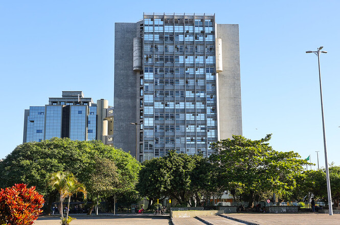 Na imagem, há um prédio de arquitetura moderna com fachada envidraçada e estrutura de concreto. O edifício é alto e cercado por árvores e vegetação. O céu está claro, indicando um dia ensolarado. Em primeiro plano, há plantas e algumas pessoas, sugerindo que a área pode ser um espaço público ou praça.