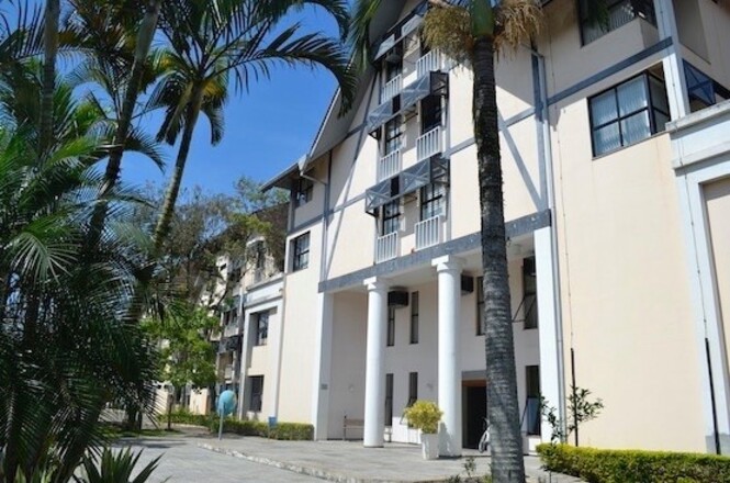 A imagem mostra um edifício de arquitetura contemporânea com janelas grandes e uma fachada clara. Há colunas destacando a entrada principal. Ao redor do edifício, há palmeiras e vegetação formando uma área ajardinada. O céu está azul, sugerindo um dia ensolarado.