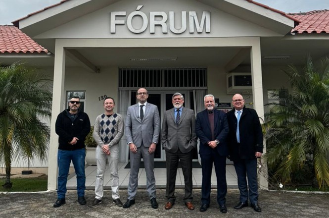 A imagem mostra seis homens em frente a um prédio com a inscrição "FÓRUM" na fachada. Eles estão em pé lado a lado, vestidos formalmente, com ternos e casacos. O edifício tem um telhado de telhas vermelhas e algumas plantas ao redor. A cena parece ser de um evento ou visita oficial.