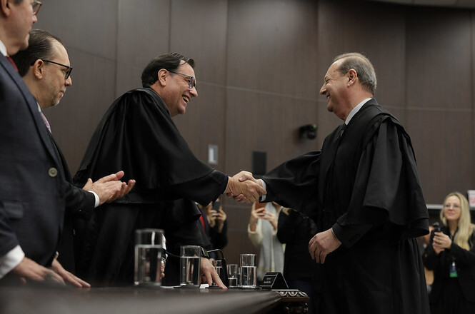 A imagem mostra homens em trajes formais, possivelmente togas, em um ambiente que parece ser uma sala de cerimônia ou tribunal. Dois homens estão no centro, cumprimentando-se com um aperto de mão e sorrindo. Outras pessoas ao redor observam e aplaudem a interação. Há copos de água sobre a mesa e uma parede de madeira ao fundo. O ambiente parece formal e solene.