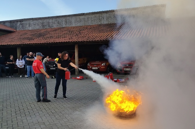 A imagem mostra um grupo de pessoas participando de uma simulação de combate a incêndio. No centro, uma mulher está usando um extintor de incêndio para apagar as chamas ressaltadas por fumaça. Ao lado dela, um instrutor está observando e orientando. Vários carros estão estacionados em segundo plano, e outras pessoas estão assistindo a demonstração. O cenário parece ser um pátio aberto com um telhado ao fundo.