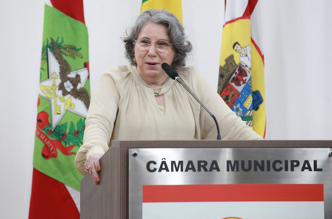 Na imagem, há uma pessoa falando em um pódio que tem a inscrição "CÂMARA MUNICIPAL". Ela está usando uma blusa clara e óculos. Atrás dela, há bandeiras coloridas, possivelmente de estados ou municípios. A configuração sugere um ambiente formal, como uma sessão legislativa ou evento oficial.