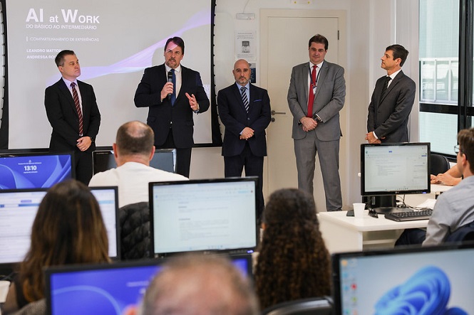 A imagem mostra um grupo de cinco homens de terno em pé na frente de uma sala, que parece ser um ambiente de treinamento ou apresentação. Eles estão em frente a uma tela de projeção que exibe o título "AI at Work: Do Básico ao Intermediário". Uma das pessoas está falando em um microfone. Na sala, há várias pessoas sentadas em frente a computadores, assistindo à apresentação. Algumas telas dos computadores mostram conteúdo relacionado à inteligência artificial.